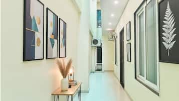 A corridor with framed artwork on the walls, a small table with a vase and a lamp, and a large window with a fern decoration.