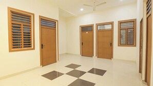 A hallway with four wooden doors, each with a window with horizontal blinds. The floor has a checkered pattern of light and dark tiles. - Fabhotel Lotus (Agra)