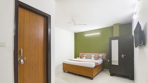 A bedroom with a wooden door, a bed with a white mattress and colorful pillows, a dark wardrobe, and a ceiling fan. - Fabhotel Raas Inn (Lucknow)