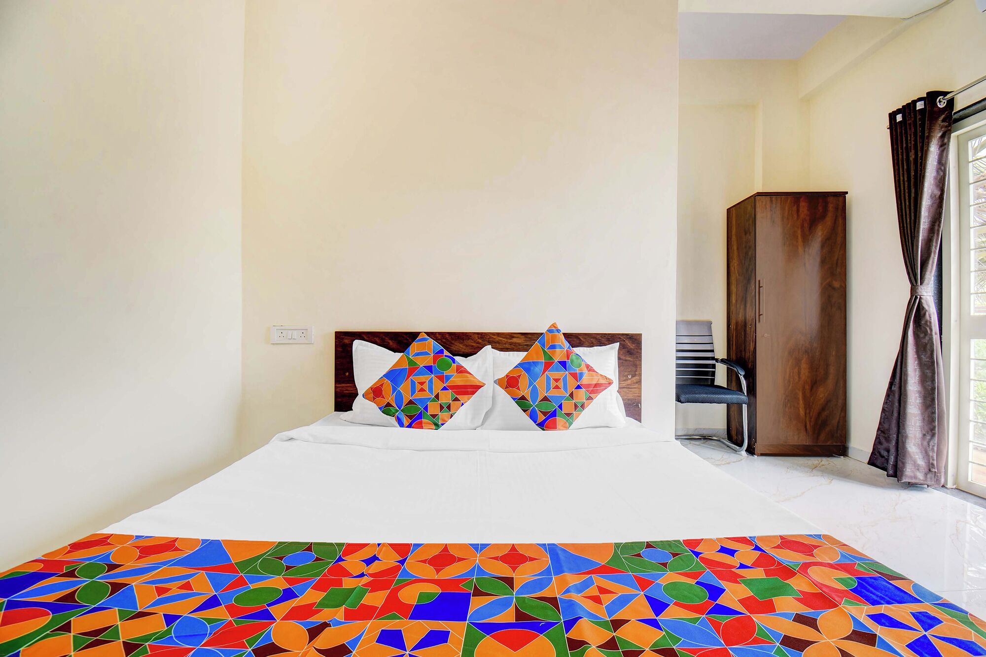 Una habitación de hotel con cama, dos almohadas, una silla, un armario y una ventana con cortinas.