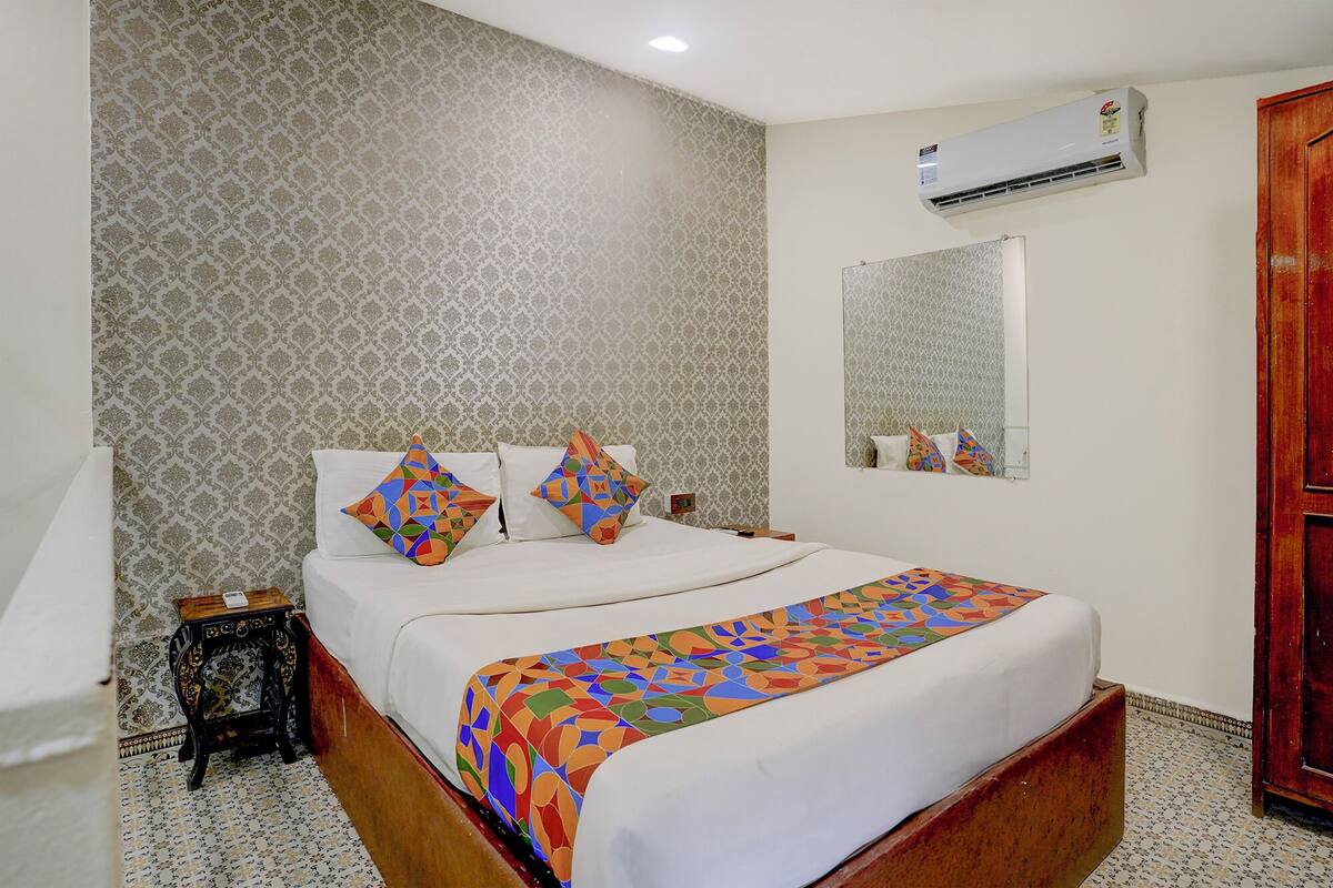 A neatly made bed with colorful pillows, a small bedside table, a wall-mounted air conditioner, and a mirror above the bed.