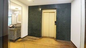 A hallway with a wooden floor, a grey sofa, a wall-mounted light fixture, and a door with a security panel. - Fabhotel Crescent (Faridabad)