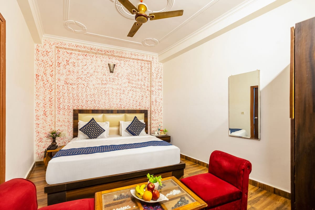 A hotel room with a bed, a red armchair, a small table with fruit, a ceiling fan, and a mirror on the wall.