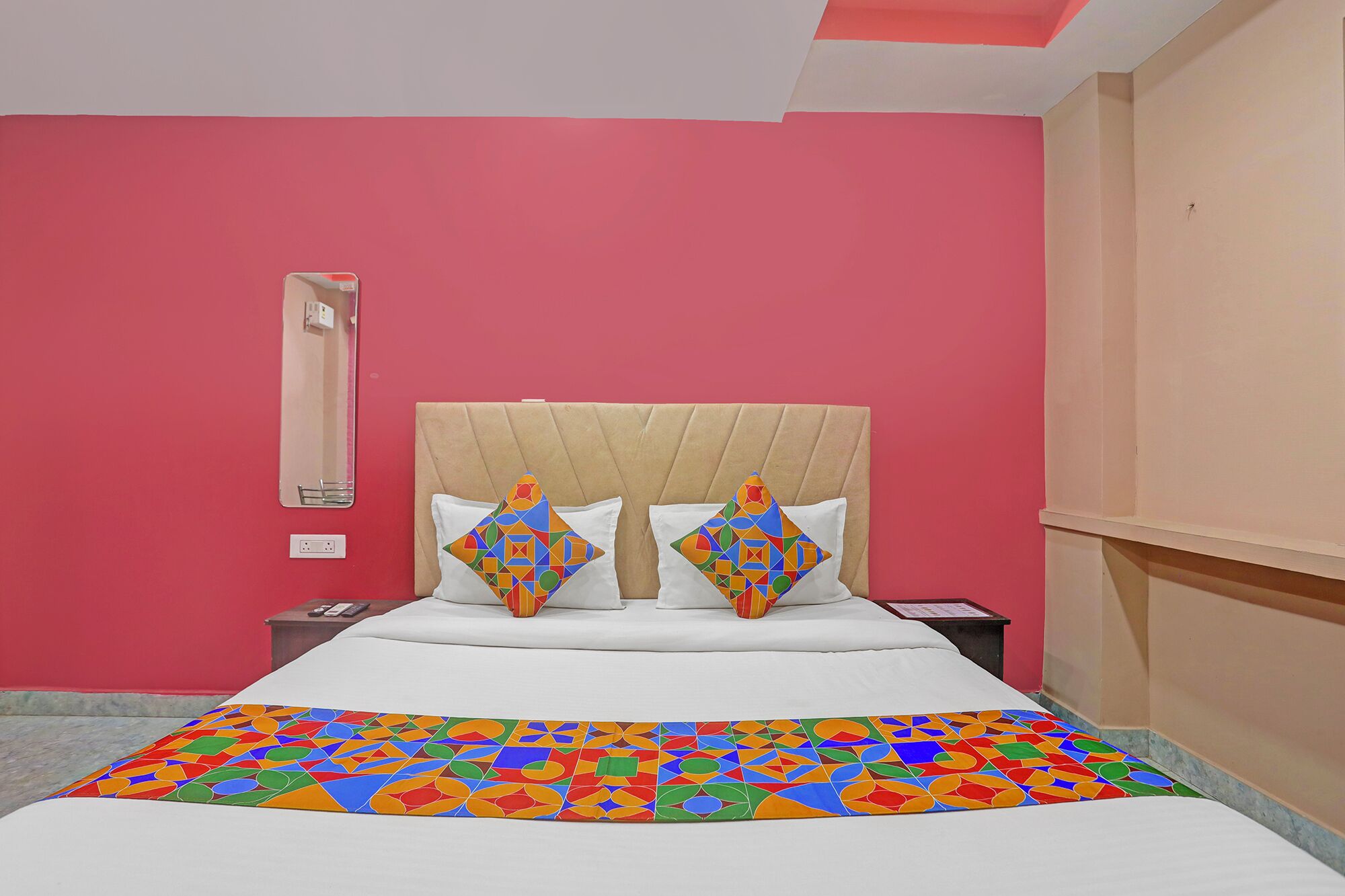 A hotel room with a pink wall, a beige headboard, and a colorful geometric-patterned bedspread.