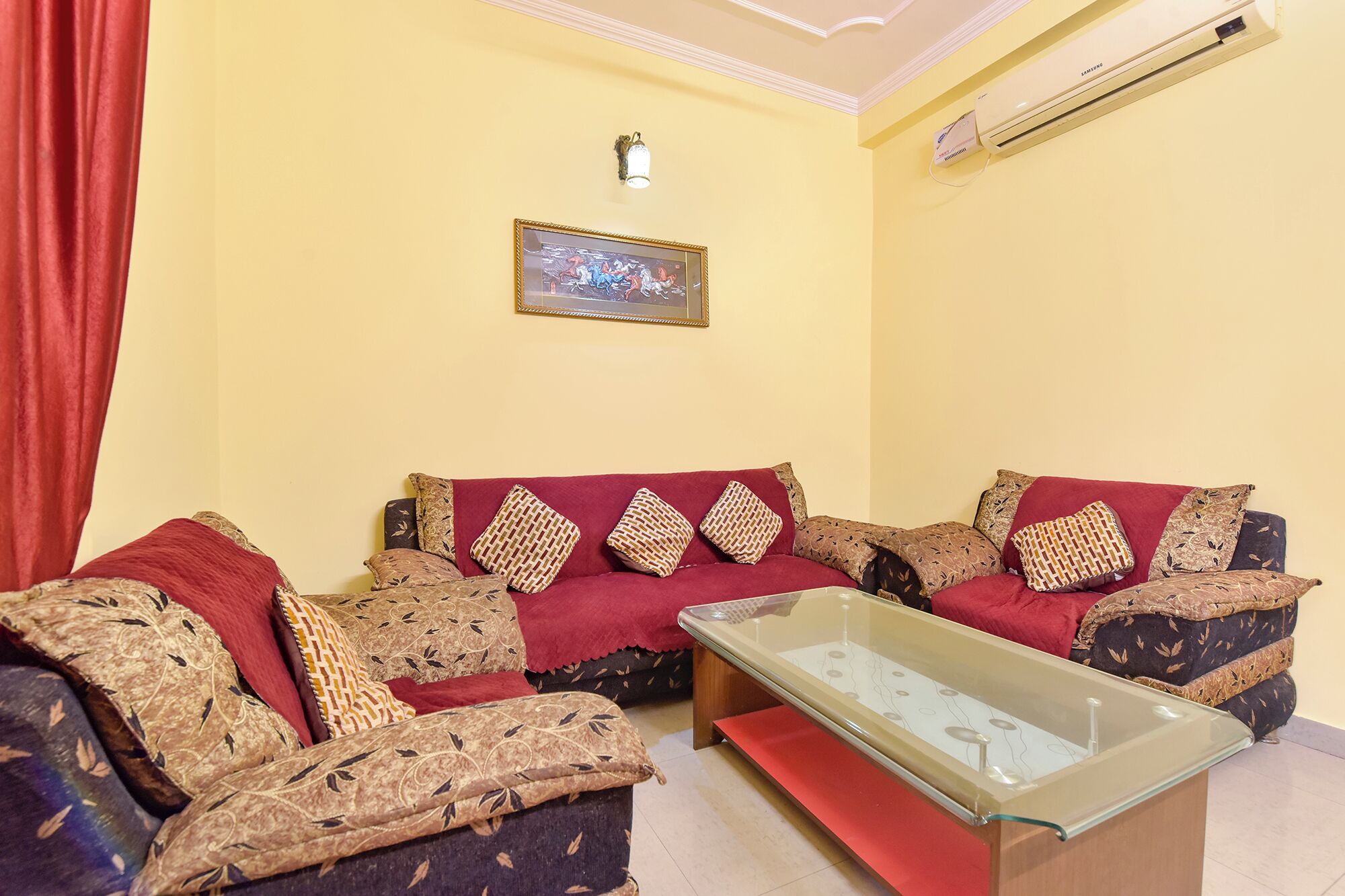 A living room with a sofa set, a glass coffee table, and a wall-mounted air conditioner.