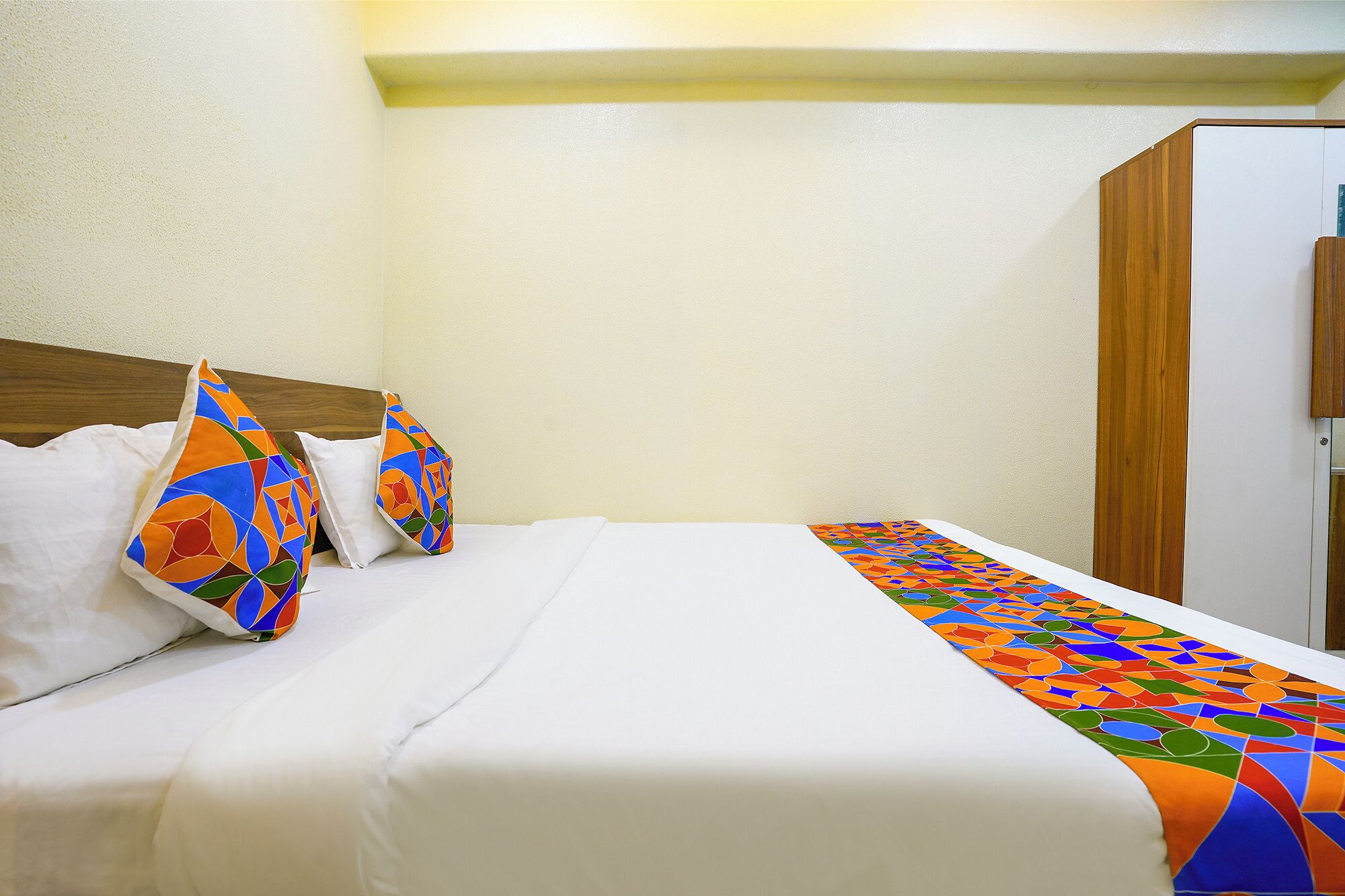 A hotel room with a bed, white linens, a colorful bedspread, and a wooden wardrobe.