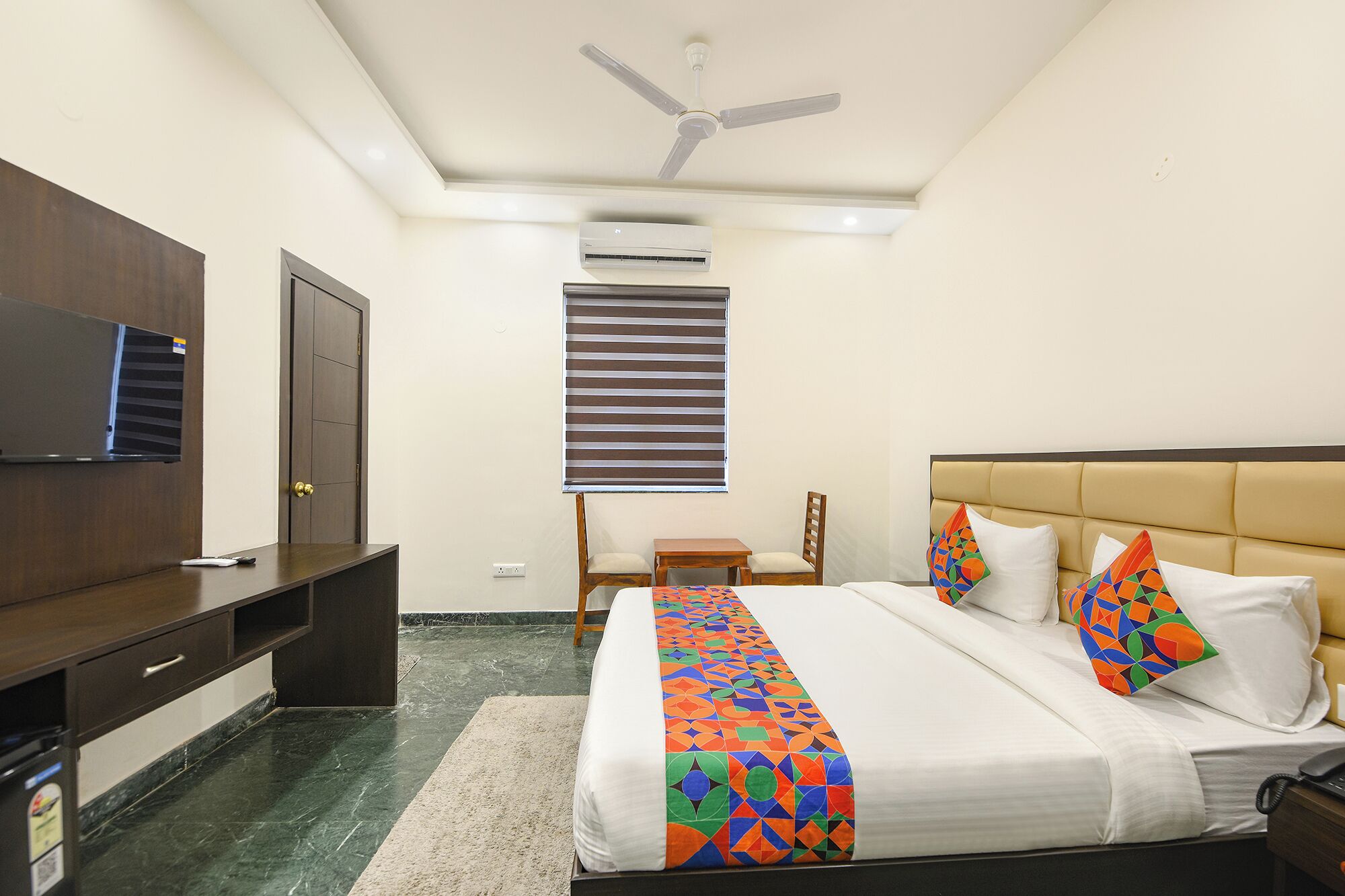 A hotel room with a bed, a desk, a chair, a television, and an air conditioning unit.