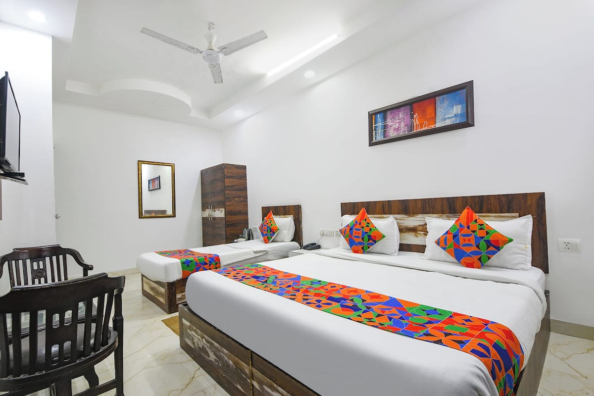 A hotel room with a large bed, two smaller beds, a wooden wardrobe, a chair, a ceiling fan, and a framed artwork on the wall.