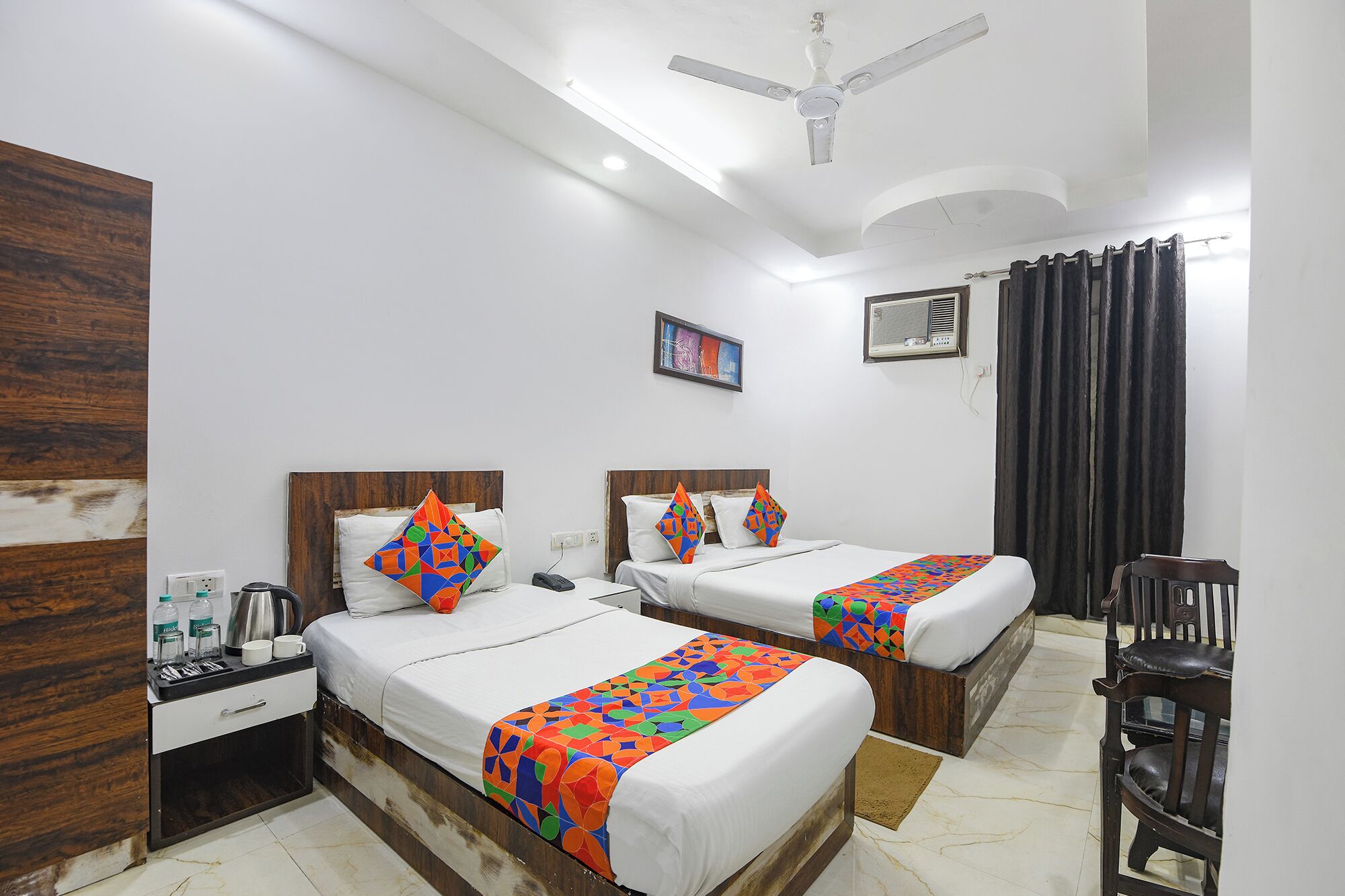 A hotel room with two beds, a ceiling fan, a painting, and a window with curtains.