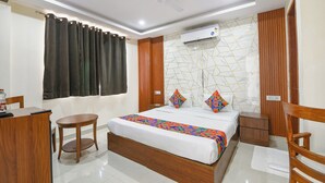 A hotel room with a bed, a desk, a chair, a small table, an air conditioning unit, and a ceiling fan. - Fabhotel Laxmi Mahal (New Delhi)