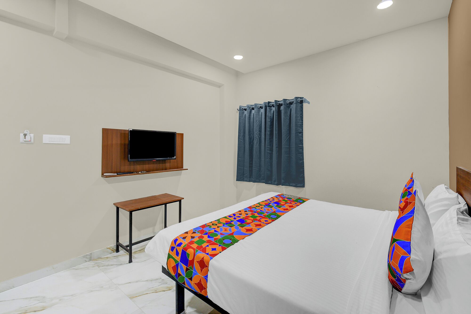 Une chambre d’hôtel avec un lit, un téléviseur fixé au mur, une petite table et une fenêtre avec des rideaux.