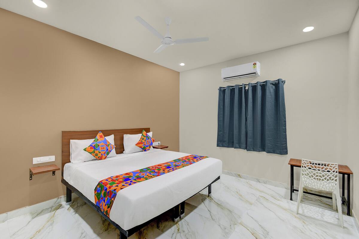 A hotel room with a bed, a ceiling fan, an air conditioning unit, a window with curtains, a small table, and a chair.