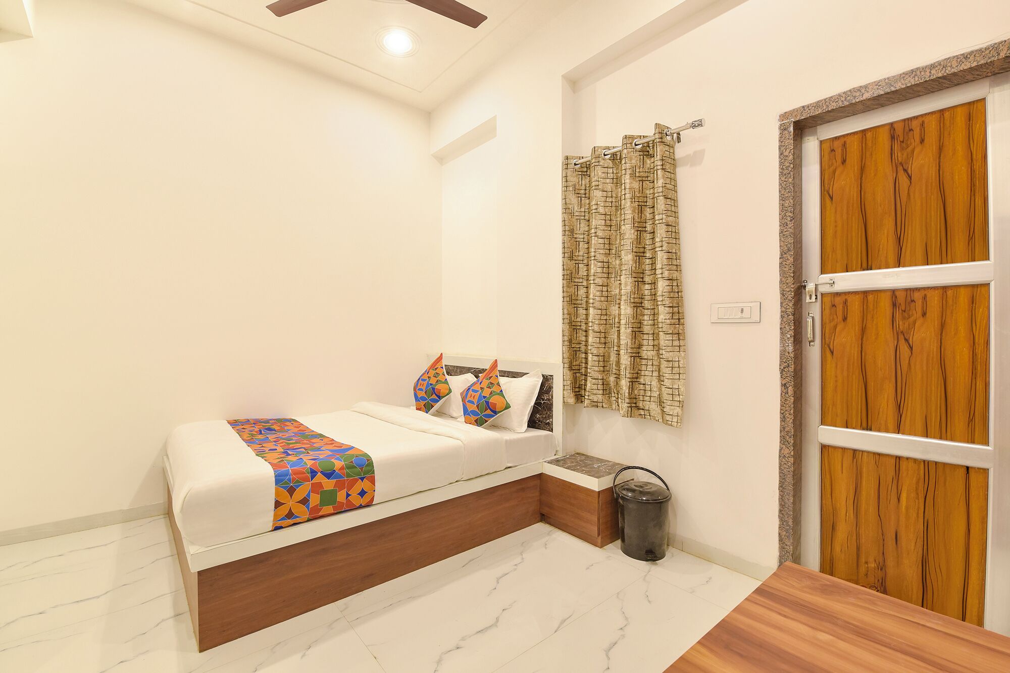 Un dormitorio con cama, una puerta de madera, una mesita y un cubo de basura.