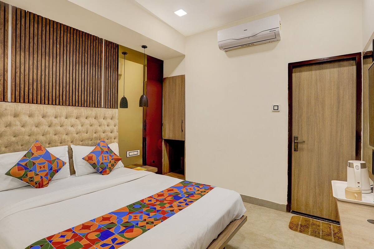A hotel room with a tufted headboard, a bed with colorful geometric pillows, a patterned runner, a wooden door, and a coffee machine on the side table.