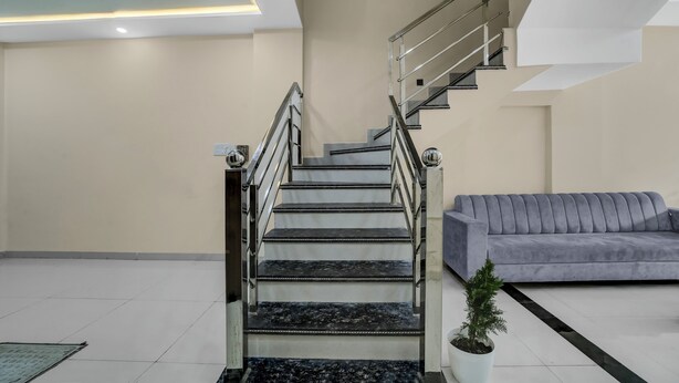 A modern staircase with metal railings, a grey sofa, and a potted plant in a spacious room with white walls and a tiled floor. - Fabhotel Shri Vinayak (Bhopal)