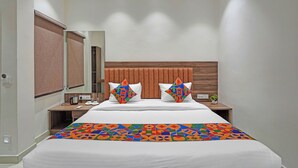 Une chambre d’hôtel moderne avec un grand lit, deux tables de chevet, une tête de lit en bois, un miroir et une couvre-lit aux motifs géométriques et aux couleurs vives.