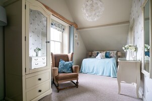 Un dormitorio con un armario grande, una silla, una cama con colcha azul y una mesita con un florero.