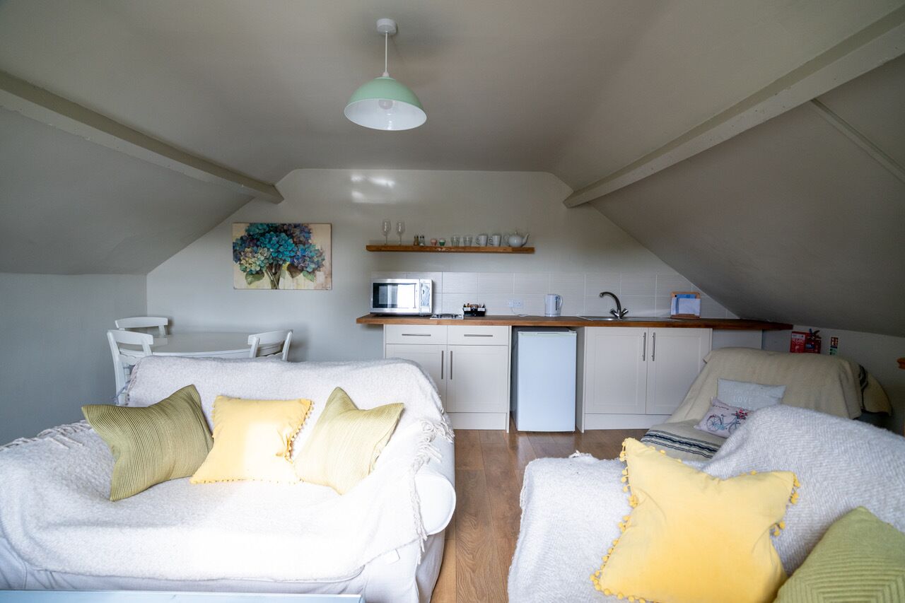 A cozy loft bedroom with a white bed, a white sofa, and a kitchenette in the background.