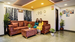 Un área de recepción con un sofá marrón, dos plantas en macetas y un reloj de pared.