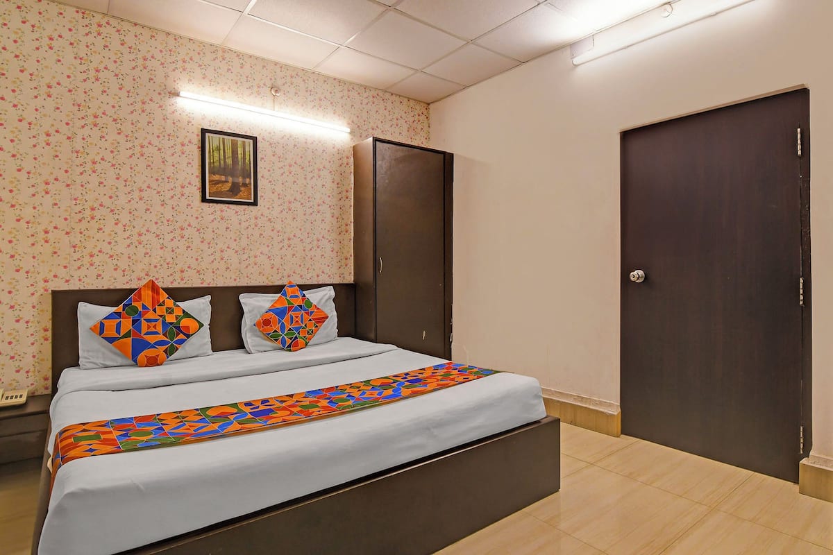 A neatly made bed with a colorful geometric-patterned bedspread and pillows, a framed picture on the wall, and two dark wooden doors.