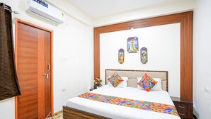 A bedroom with a wooden bed frame, a white bedspread, and colorful geometric pillows. There is a wall-mounted air conditioner and a wooden door with a geometric pattern. - Fabhotel Sp 97 (Indore)