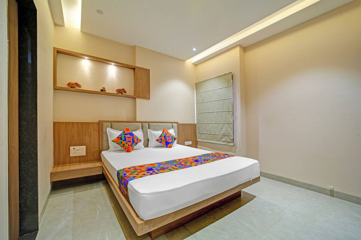 A modern hotel room with a large bed, a wooden headboard, a nightstand, a wall-mounted shelf with decorative items, and a window with a view.