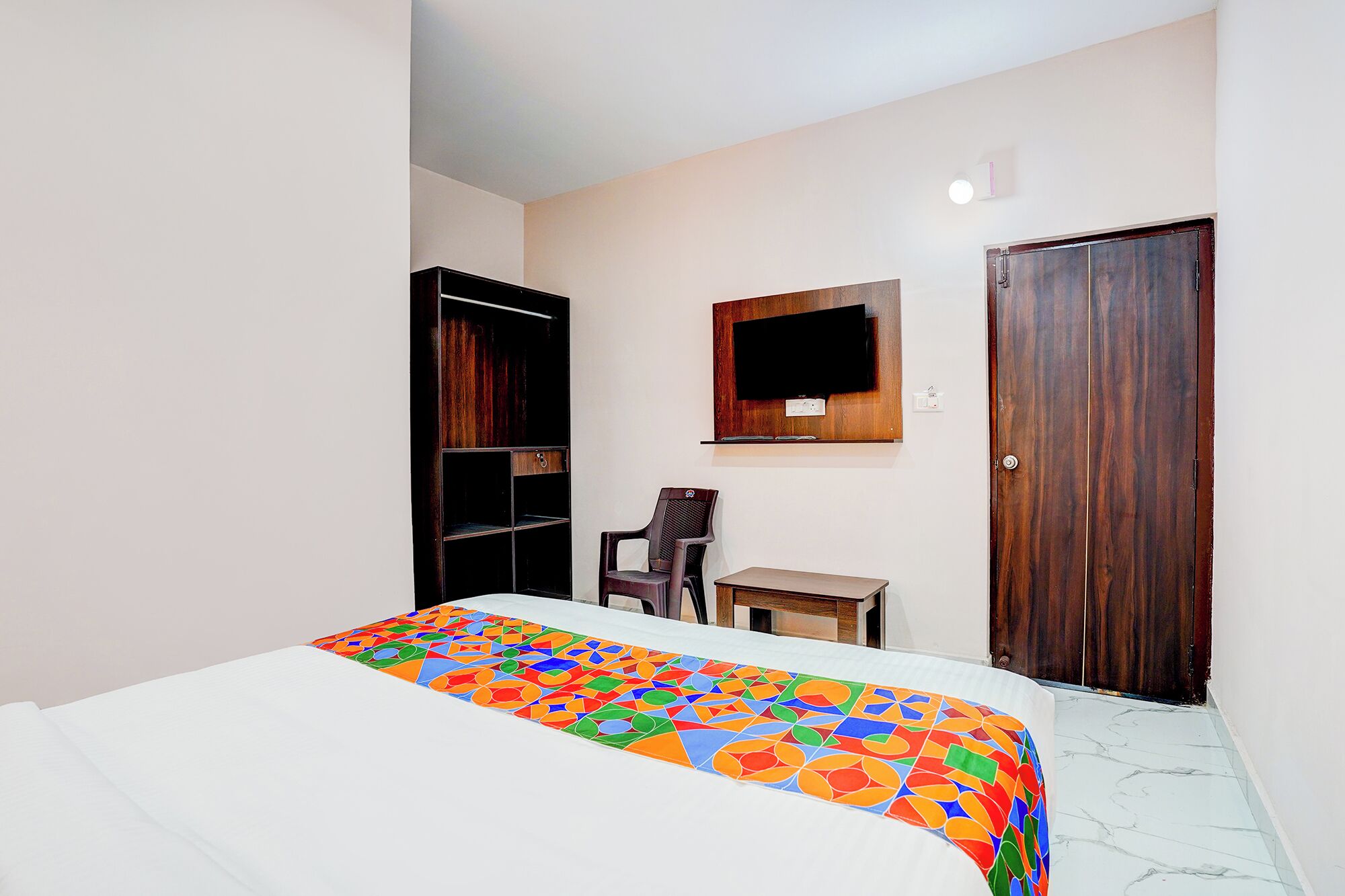 A bedroom with a bed, a TV mounted on the wall, a wooden wardrobe, a small table, and a chair.