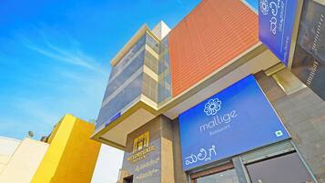 A hotel with a modern facade, a visible sign for 'MYSORE GATE HOTEL', and a prominent 'malige' restaurant sign.