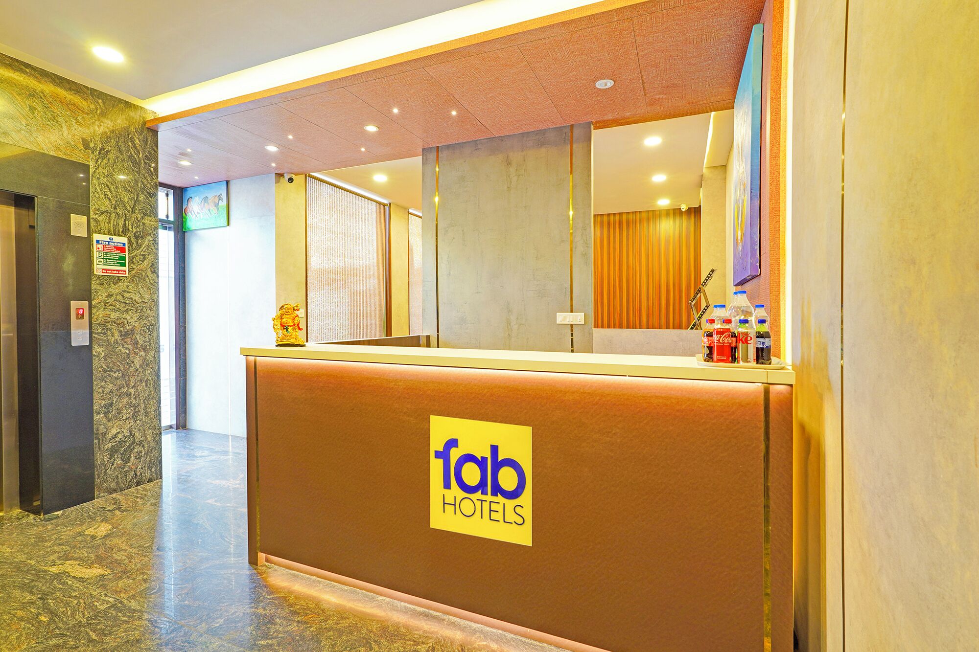 Een receptiebalie met een 'Fab Hotels' bord, een brandmeldpaneel en een Coca-Cola koelkast.