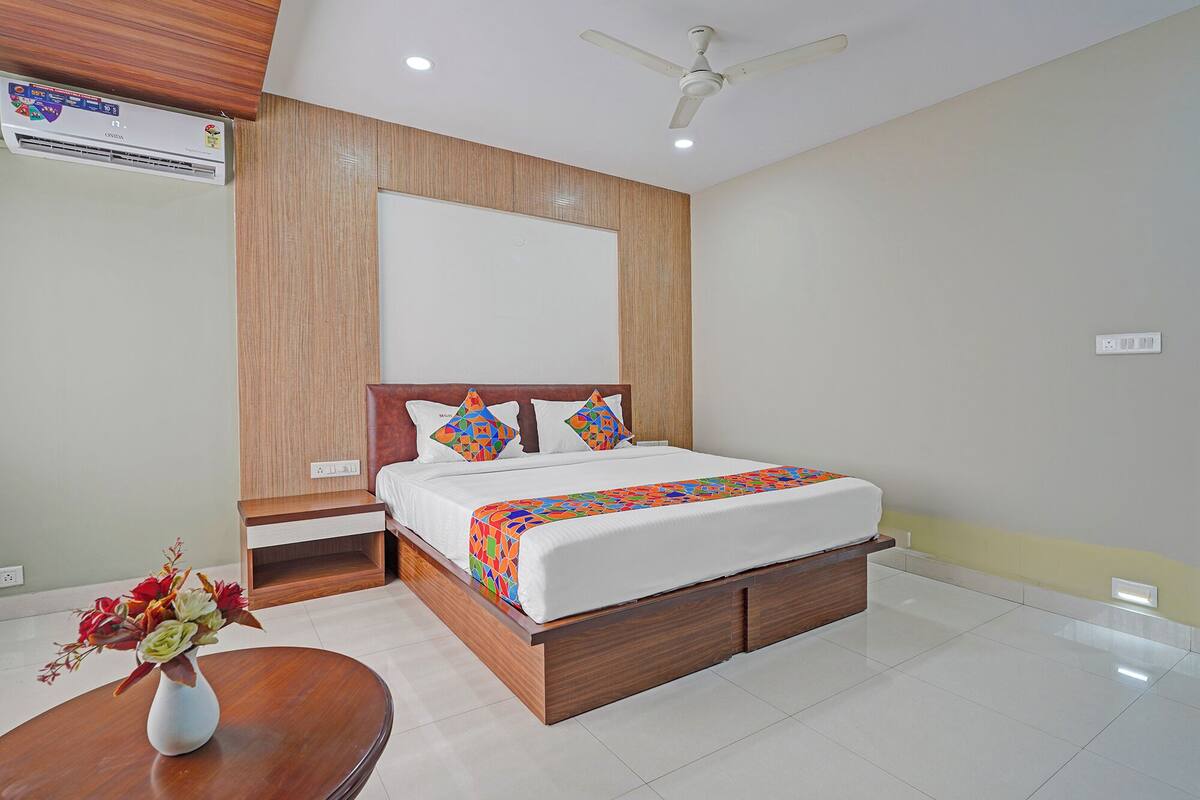 A modern hotel room with a wooden headboard, a large bed with colorful bedding, a bedside table, and a small round table with a vase of flowers.