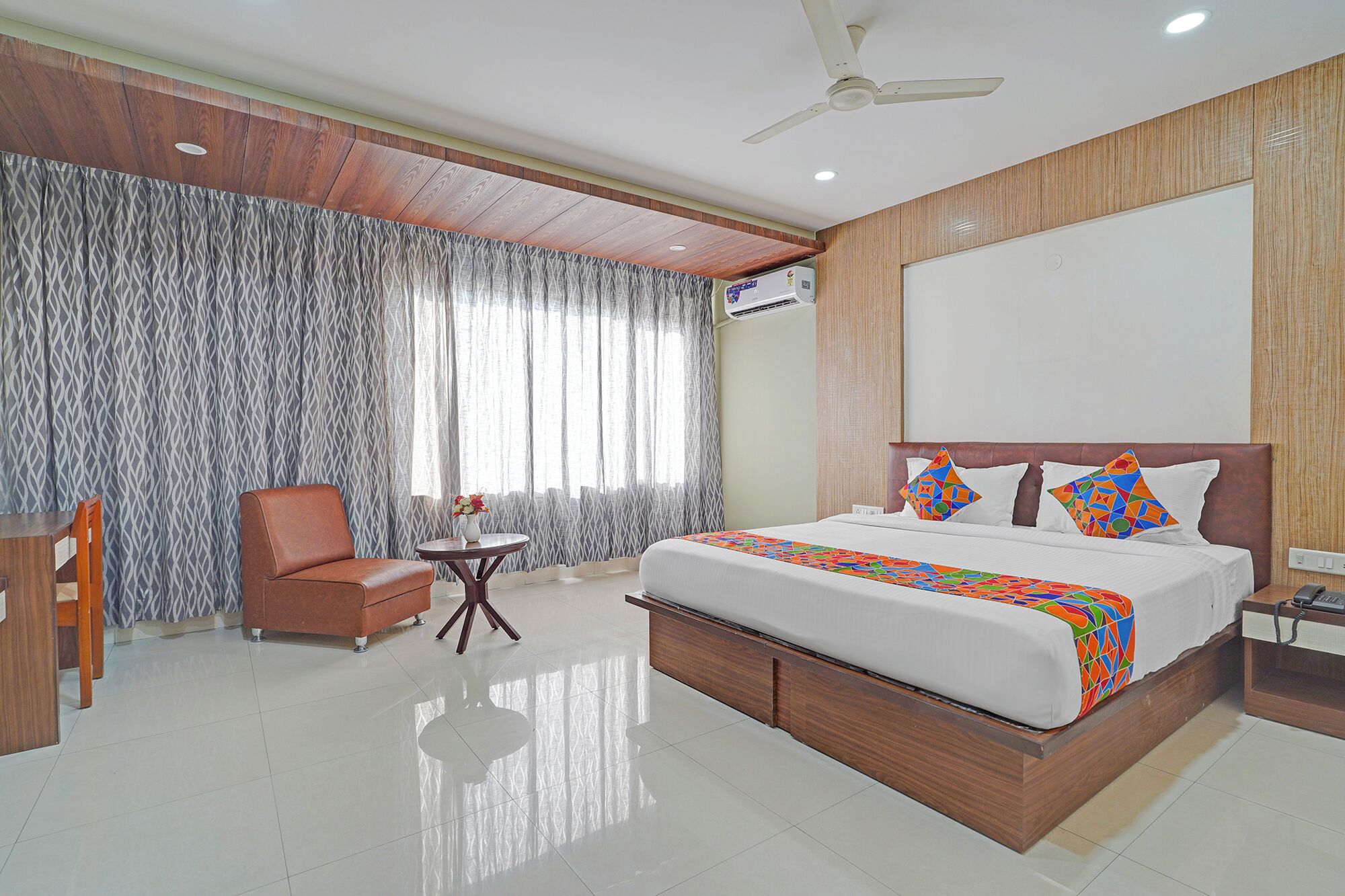 A hotel room with a large bed, a wooden headboard, a ceiling fan, and a window with patterned curtains.