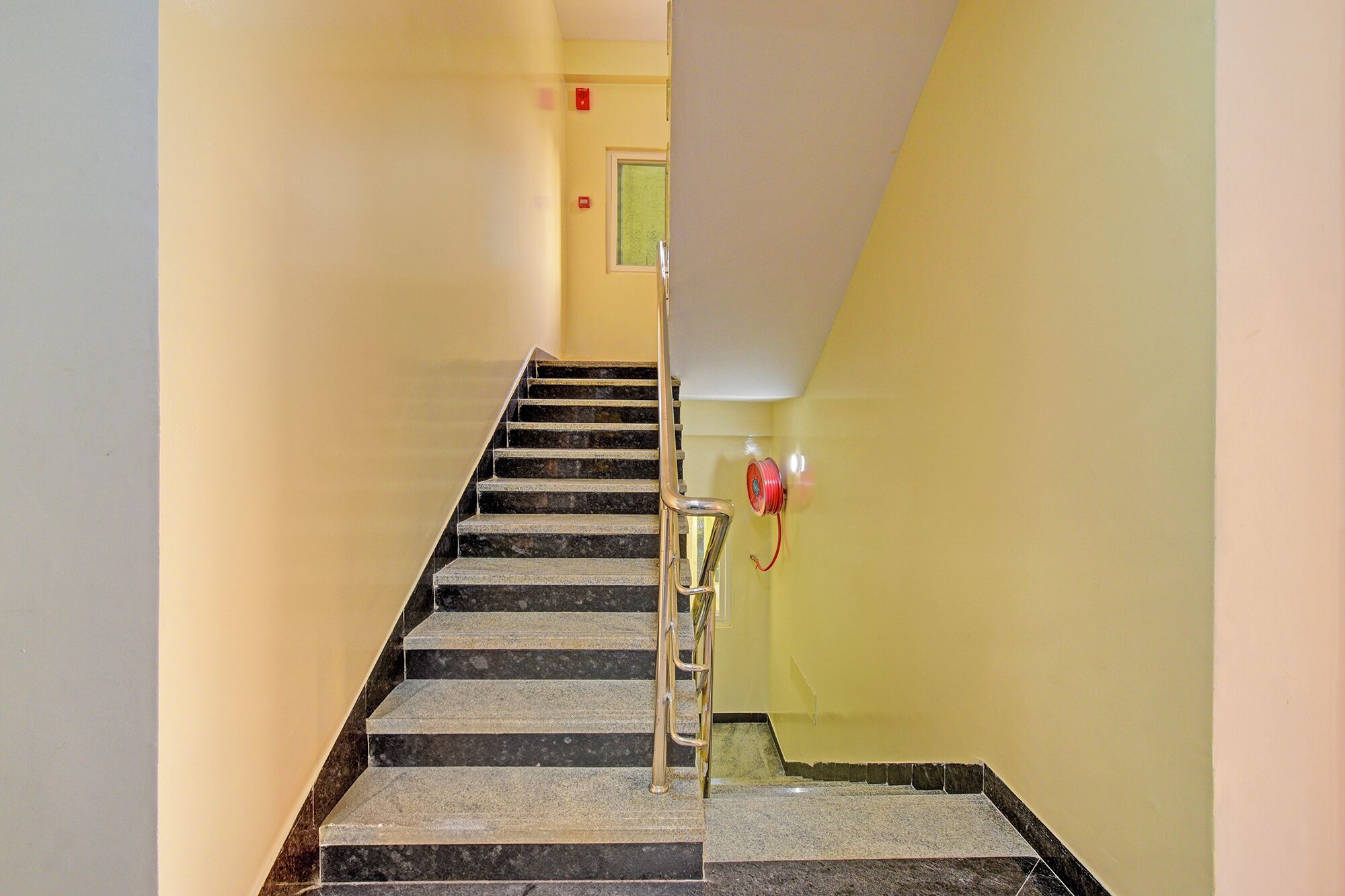 Un escalier avec des marches en marbre et une rampe métallique, menant à une pièce dont un mur est jaune et qui possède une fenêtre.