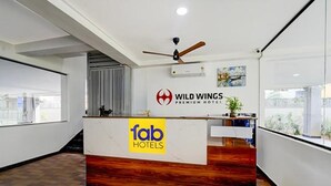 Un mostrador de recepción con detalles de madera y un cartel de 'fab hotels'. Se ve un ventilador de techo sobre el mostrador, y un cartel de 'Wild Wings Premium Hotel' en la pared.