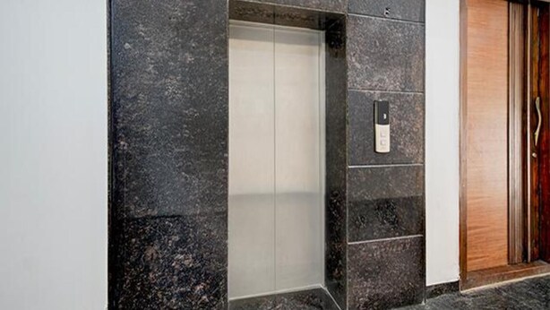 Un ascensor moderno con puerta metálica, ubicado en un edificio con paredes de piedra oscura y una puerta de madera a la derecha.
