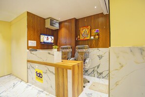 A hotel reception area with a wooden reception desk, two massage chairs, a television, and a check-out sign. - Fabhotel Shri Shubh Palace (Indore)