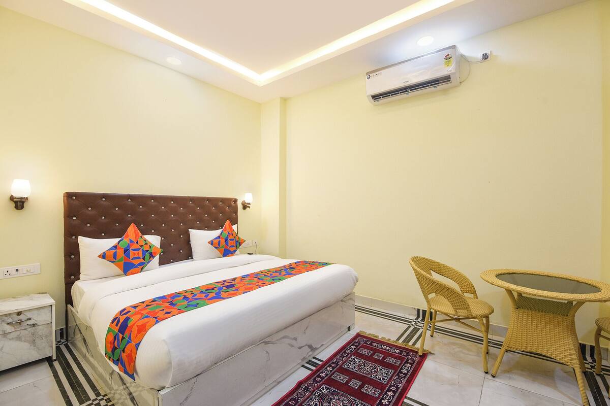 A hotel room with a bed, a small table, two chairs, an air conditioning unit, and a wall-mounted light fixture.