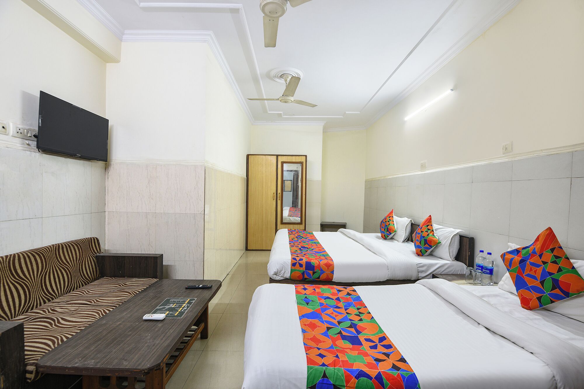 Habitación de hotel con sofá, una mesa, dos camas con almohadas coloridas, televisión y ventilador de techo.