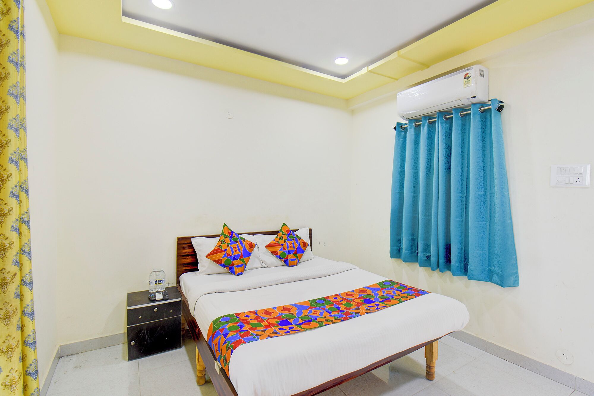 A bedroom with a bed, two pillows, a bedside table, a water bottle, and a window with curtains.