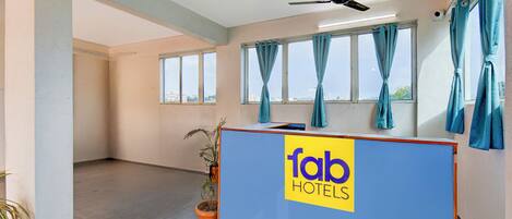 Uma área de recepção de hotel com um balcão azul, um ventilador de teto e janelas com cortinas azuis.
