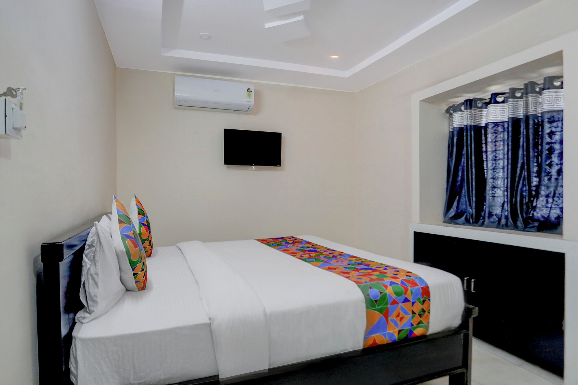 A hotel room with a bed, a flat-screen TV mounted on the wall, and a window with patterned curtains.