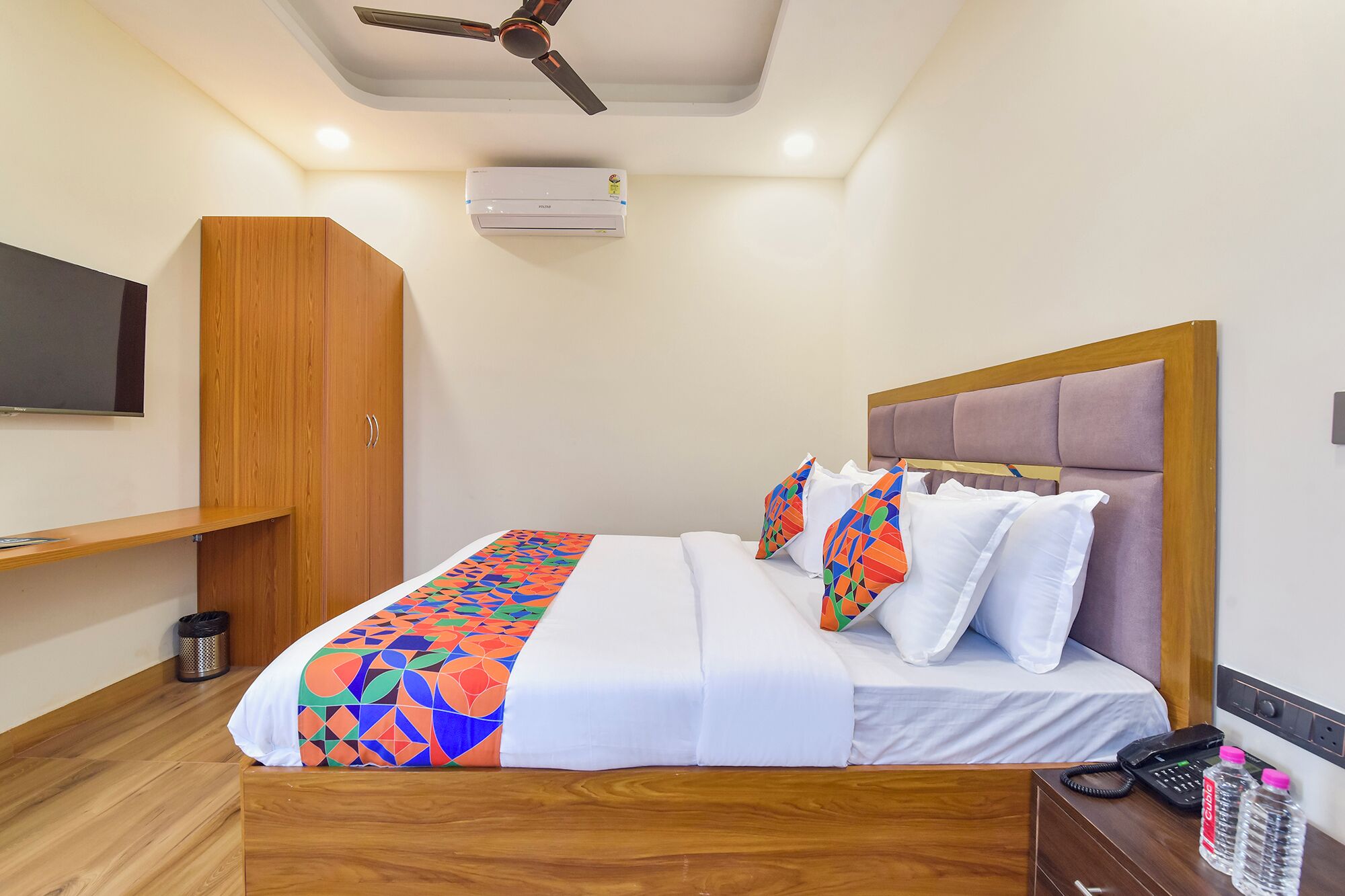 A hotel room with a bed, wooden wardrobe, ceiling fan, air conditioning unit, bedside table with a phone and water bottles, and a TV mounted on the wall.