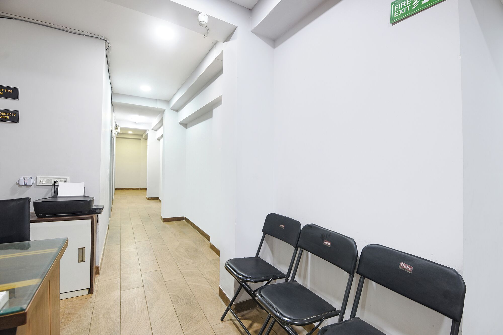 A hallway with white walls, a fire exit sign, and a row of black folding chairs.
