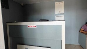A reception desk with a 'RECEPTION' sign, a white door, and a person sitting on a bench in the background. - Fabhotel Ace Executive (Pune)