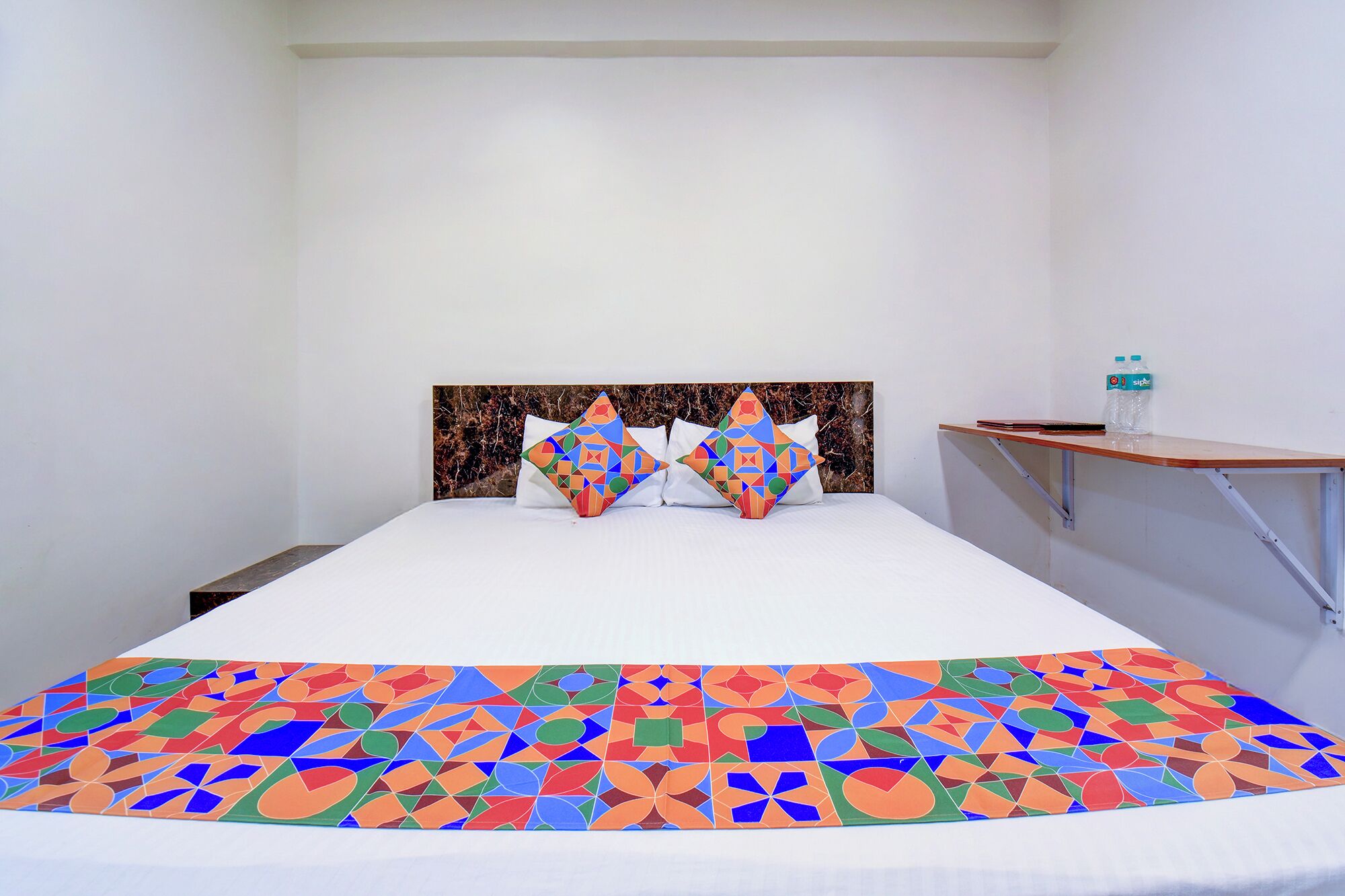 A bed with a patterned bedspread, two pillows with geometric designs, a headboard with a marble-like texture, and a small table with a water bottle.