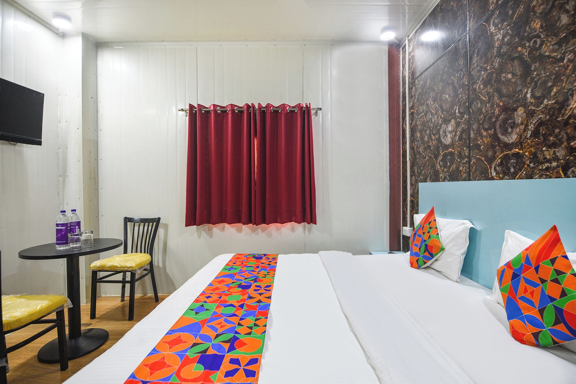 Una habitación de hotel con cama, una mesita con sillas, televisión y ventana con cortinas rojas.