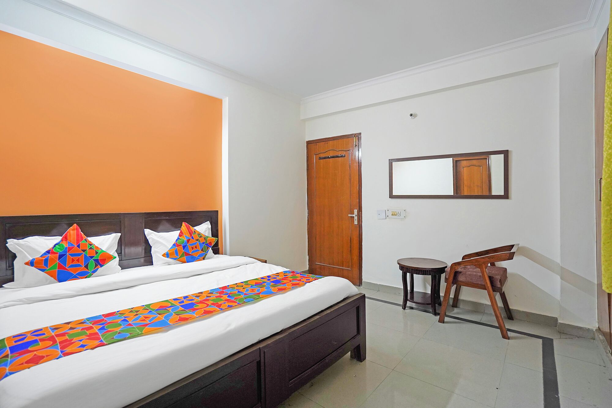A hotel room with a bed, a chair, a small table, and a mirror on the wall.