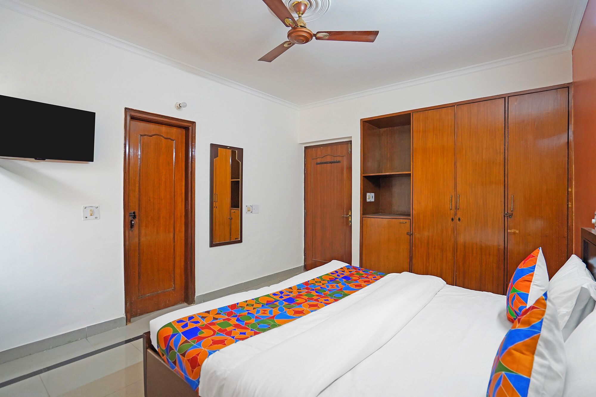 A bedroom with a bed, a ceiling fan, wooden wardrobes, a TV mounted on the wall, and a mirror.