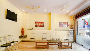 A hotel room with a TV, ceiling fans, a painting, a table, chairs, and a view through the window. - Via Ashir Inn (Mumbai)