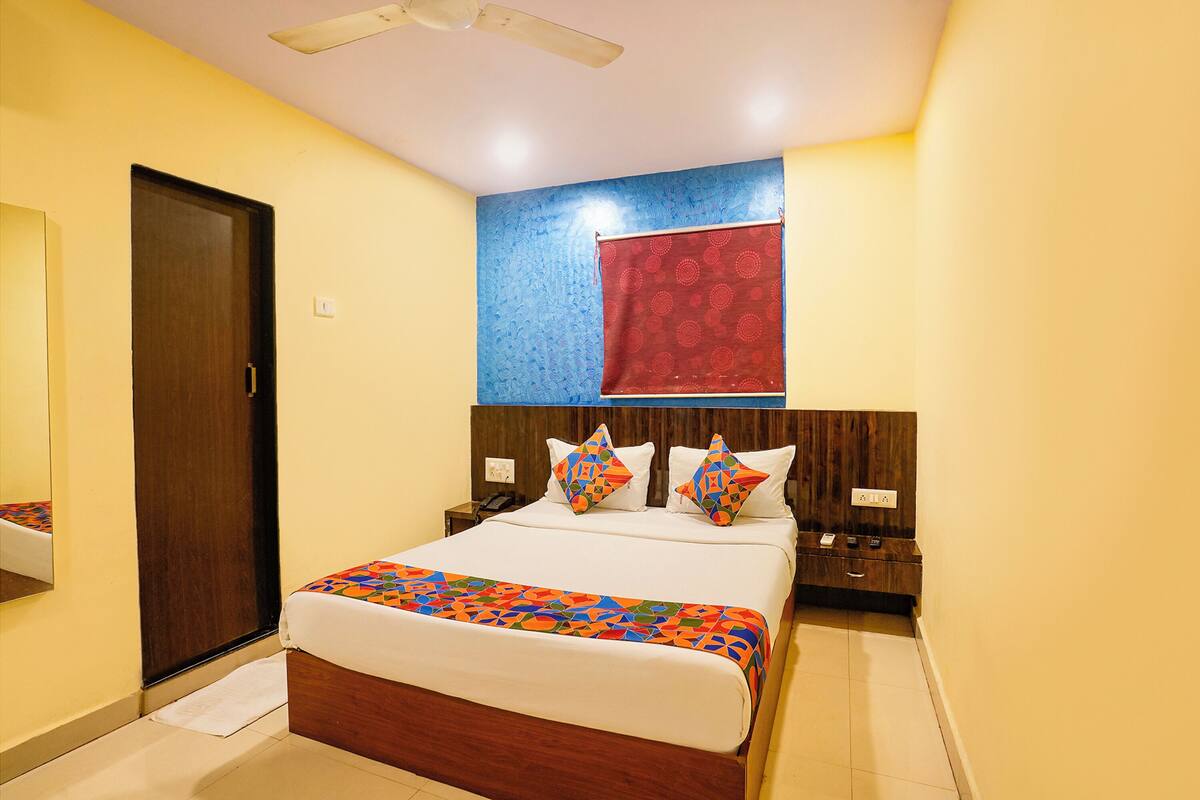 A hotel room with a bed, a wooden headboard, a nightstand, a mirror, and a red patterned curtain.
