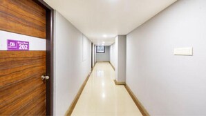 A hallway with a wooden door on the left, a light-colored floor, and a plain wall with a light switch. - Fabhotel D Grand (Madurai)